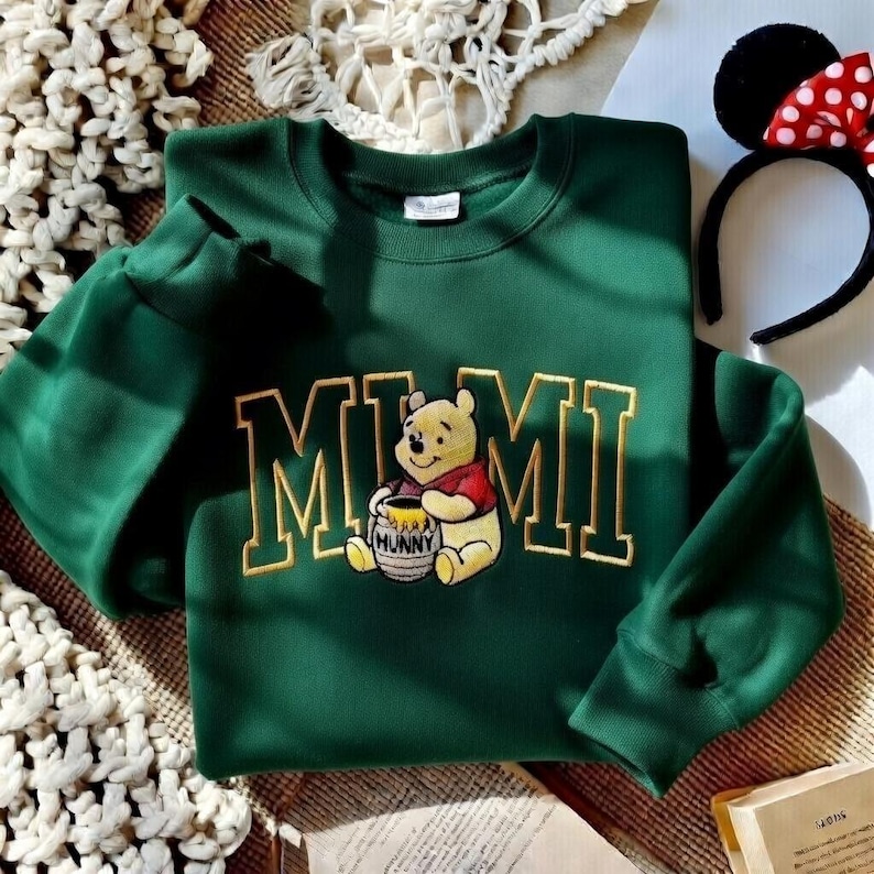 以下が含まれることがあります： 金色の文字で「MIMI」と刺繍された、ダークグリーンのスウェットシャツ。ハチミツの壺を持ったくまのプーさんのグラフィックも描かれています。赤と白のドット柄のリボンが付いた黒いヘアバンドも見えます。