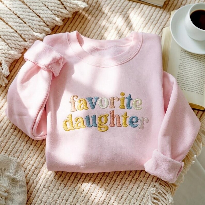 May include: A pale pink sweatshirt featuring the embroidered words "favorite daughter" in pastel shades. The garment has a crew neck and rolled-up sleeves. The image is captured from above.