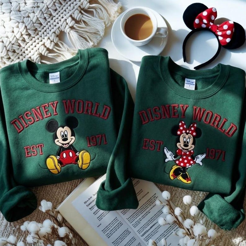 May include: Two green sweatshirts with embroidered "Disney World" and "EST 1971" text. One features Mickey Mouse, the other Minnie Mouse. A Minnie Mouse ear headband and a cup of coffee are also visible.