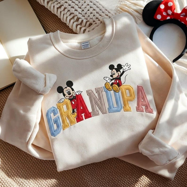 May include: Cream-coloured sweatshirt featuring the word "GRANDPA" embroidered in colourful letters. Two Mickey Mouse cartoon characters are above the letters. A Minnie Mouse headband with a red bow is visible in the background.