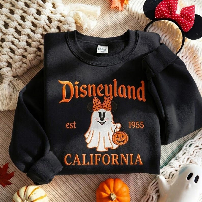 May include: A black crewneck sweatshirt featuring an embroidered graphic of a ghost wearing a polka-dot bow, holding a pumpkin bucket. The text reads Disneyland, est 1955, and California in orange lettering. The design includes a small ghost figure.