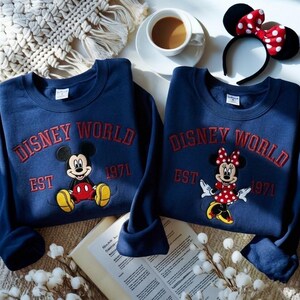 May include: Two navy blue sweatshirts with red embroidered "Disney World" text and character graphics. One features Mickey Mouse, the other Minnie Mouse. A Minnie Mouse ear headband and a cup of coffee are also visible.
