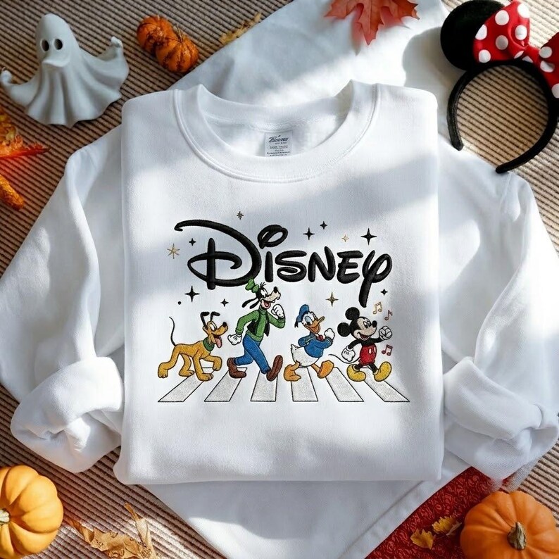 Peut inclure: Un sweat-shirt blanc &agrave; col rond avec le logo Disney brod&eacute; en noir au-dessus d'une illustration de Pluto, Dingo, Donald et Mickey marchant sur un passage pi&eacute;ton. Le motif comprend de petites &eacute;toiles dor&eacute;es et des notes de musique.