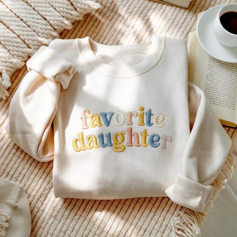 May include: A cream-coloured sweatshirt featuring the embroidered phrase "favorite daughter" in pastel shades. The long-sleeved top is neatly folded. A coffee cup and book are visible in the background.