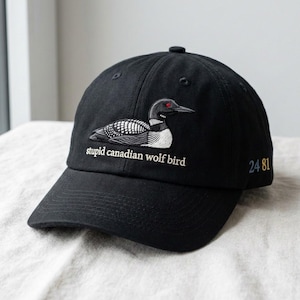 May include: Black baseball cap with an embroidered design of a loon bird and the text "stupid canadian wolf bird". The cap also features the numbers "24 81" embroidered on the side. The cap is made of a soft material.