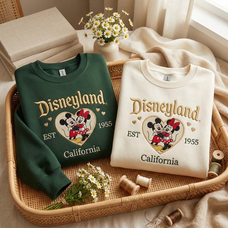May include: Two sweatshirts, one dark green and one cream, with the Disneyland logo embroidered in gold. The logo includes Mickey and Minnie Mouse in a heart, with "EST 1955 California" below. The sweatshirts are displayed in a woven tray.