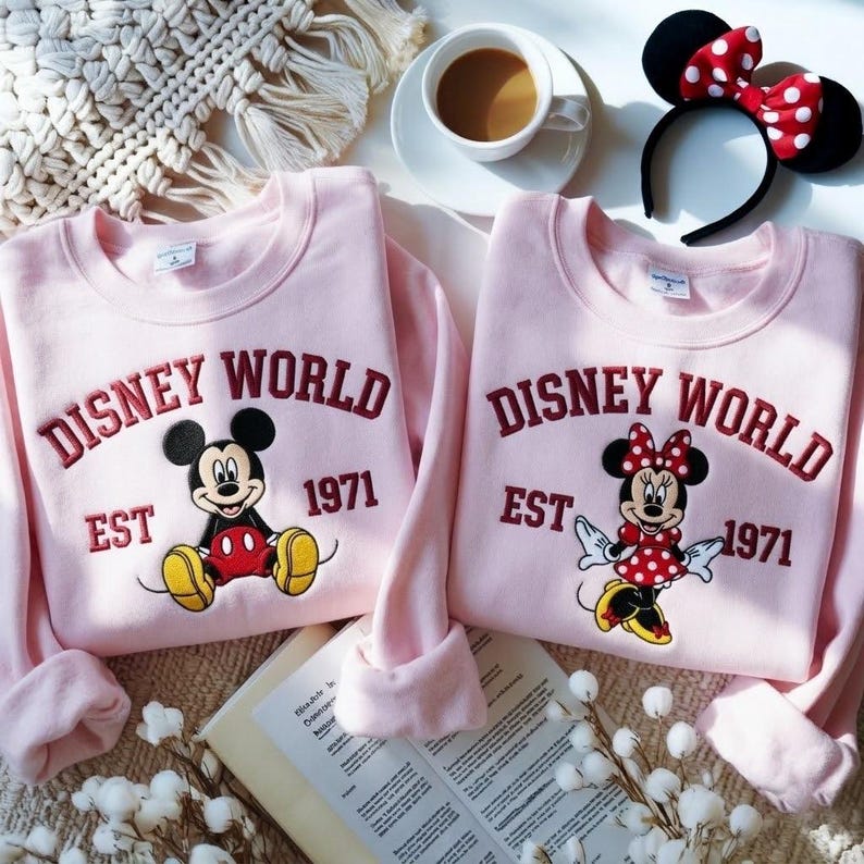 May include: Two light pink sweatshirts with the words "DISNEY WORLD EST 1971" embroidered in burgundy. One sweatshirt features Mickey Mouse, the other Minnie Mouse. A Minnie Mouse ear headband and a cup of coffee are also visible.