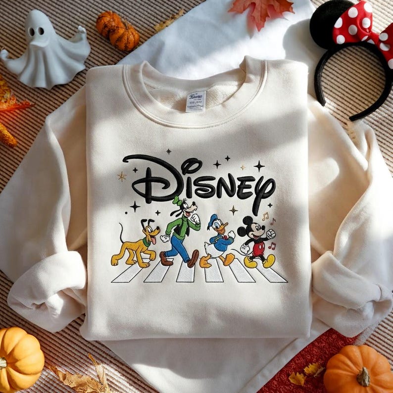 Peut inclure: Un sweat-shirt &agrave; col rond couleur cr&egrave;me arborant un logo Disney brod&eacute; en noir au-dessus d'un motif repr&eacute;sentant Pluto, Dingo, Donald et Mickey marchant sur un passage pi&eacute;ton blanc, agr&eacute;ment&eacute; de petites &eacute;toiles et notes de musique noires.