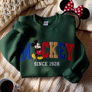 May include: A dark green sweatshirt with the word "Mickey" in blue, red, and gold lettering, and a cartoon Mickey Mouse graphic. The text "SINCE 1928" is below the word "Mickey". A pair of Mickey Mouse ears with a red and white polka dot bow is in the background.