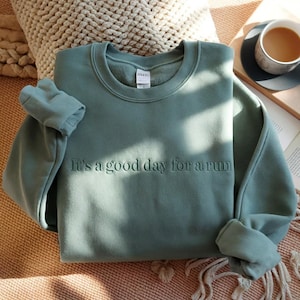 May include: A sage green crewneck sweatshirt featuring the embroidered text It's a good day for a run across the chest. The garment has long sleeves with ribbed cuffs and a ribbed neckline, showcasing a clean, solid-coloured design throughout.