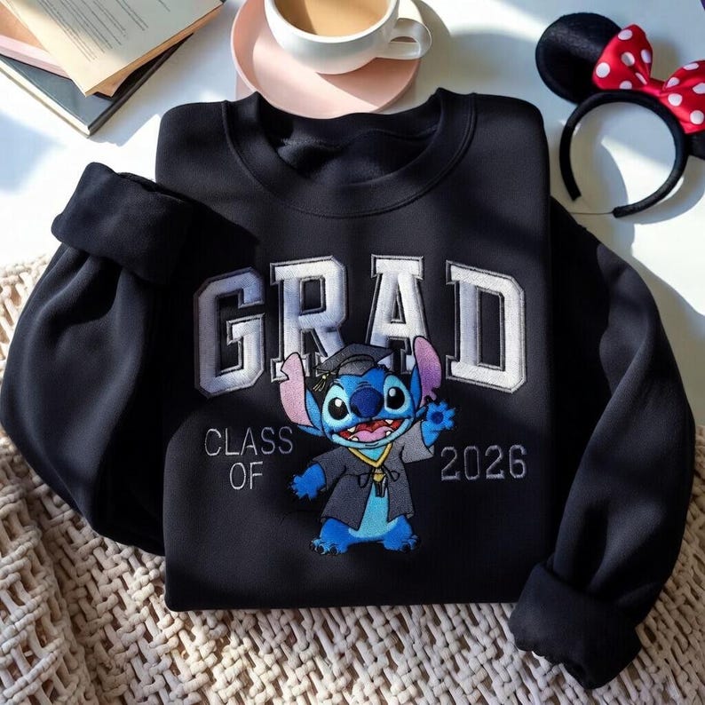 Puede incluir: Sudadera negra con la palabra "GRAD" en letras blancas. Debajo, un personaje de dibujos animados con birrete y toga de graduaci&oacute;n. El texto "CLASS OF 2026" tambi&eacute;n est&aacute; en la sudadera.