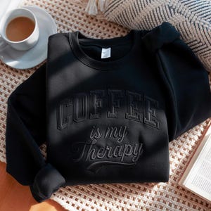 May include: White ceramic mug with a black handle, featuring the text "But First, Coffee" in black. The mug's design is simple, set against a blurred background.
