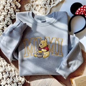 以下が含まれることがあります： 金色の文字で「MIMI」と刺繍されたライトグレーのスウェットシャツと、くまのプーさんの漫画の画像。赤と白のドット柄のリボンが付いた黒いヘアバンドも見えます。