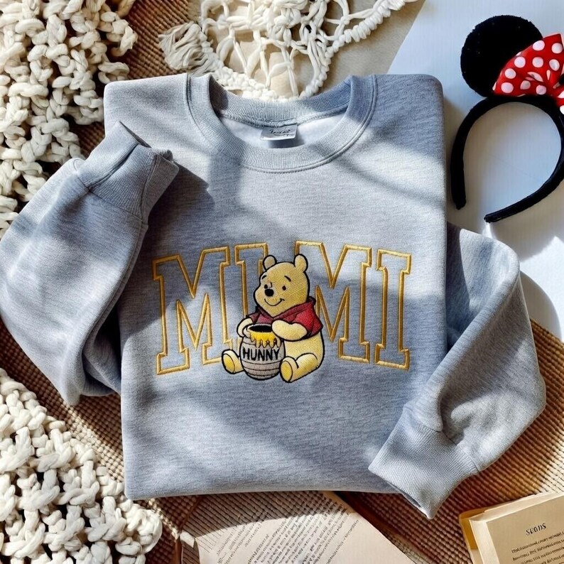 以下が含まれることがあります： 金色の文字で「MIMI」と刺繍されたライトグレーのスウェットシャツと、くまのプーさんの漫画の画像。赤と白のドット柄のリボンが付いた黒いヘアバンドも見えます。