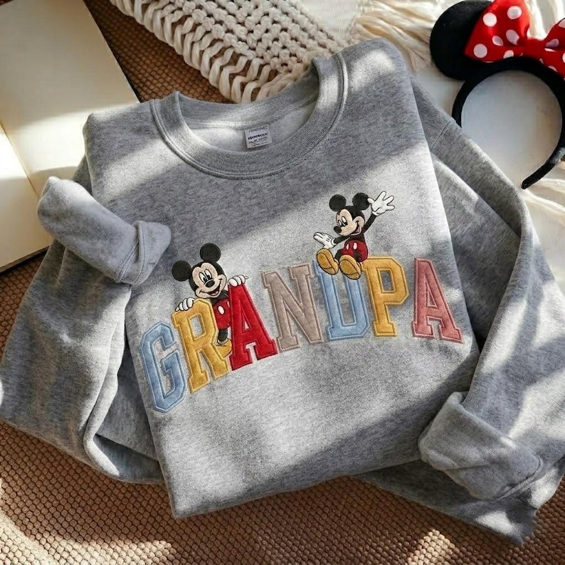 May include: A grey sweatshirt featuring the word "GRANDPA" embroidered in colourful letters. Mickey Mouse is depicted on the sweatshirt. A Minnie Mouse headband with a red bow is visible in the background.