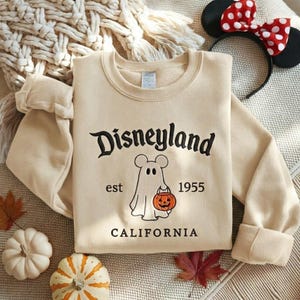Pode incluir: Uma camisola de gola redonda bege com texto bordado a preto que diz Disneyland e California. No centro, um fantasma branco com orelhas de rato segura uma ab&oacute;bora laranja, ladeado pelo texto est 1955 bordado a preto.