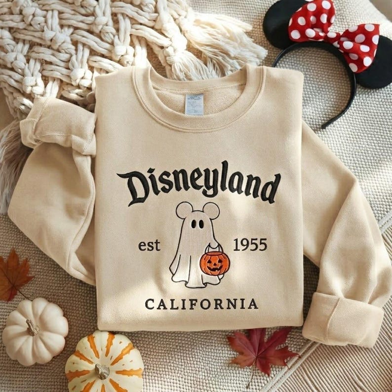 Pode incluir: Uma camisola de gola redonda bege com texto bordado a preto que diz Disneyland e California. No centro, um fantasma branco com orelhas de rato segura uma ab&oacute;bora laranja, ladeado pelo texto est 1955 bordado a preto.