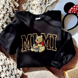 以下が含まれることがあります： 金色のブロック体で「MIMI」と書かれた黒いスウェットシャツ。テキストの下には、蜂蜜の壺を持っている漫画のくまのプーさんのキャラクターが刺繍されています。ミニーマウスの耳と赤い水玉模様のリボンが付いた黒いヘアバンドも見えます。