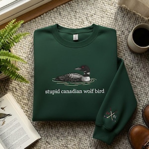 May include: A dark green sweatshirt with a graphic of a loon bird on water and the text "stupid canadian wolf bird". The sleeve has an embroidered design of crossed hockey sticks and the numbers 24 and 81.