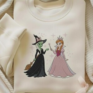 May include: Cream-coloured sweatshirt with an embroidered design of a witch and a princess. The witch has green skin, a black dress, and a broom. The princess wears a pink dress and a crown. The design is surrounded by star accents.