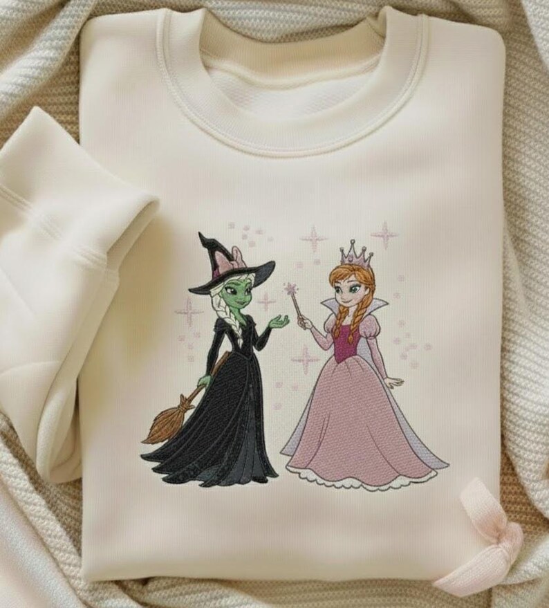 May include: Cream-coloured sweatshirt with an embroidered design of a witch and a princess. The witch has green skin, a black dress, and a broom. The princess wears a pink dress and a crown. The design is surrounded by star accents.