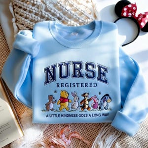 May include: Light blue sweatshirt with the word "NURSE" embroidered in navy blue, with "REGISTERED" below. Features cartoon characters and the phrase "A LITTLE KINDNESS GOES A LONG WAY". A Minnie Mouse ear headband is in the background.