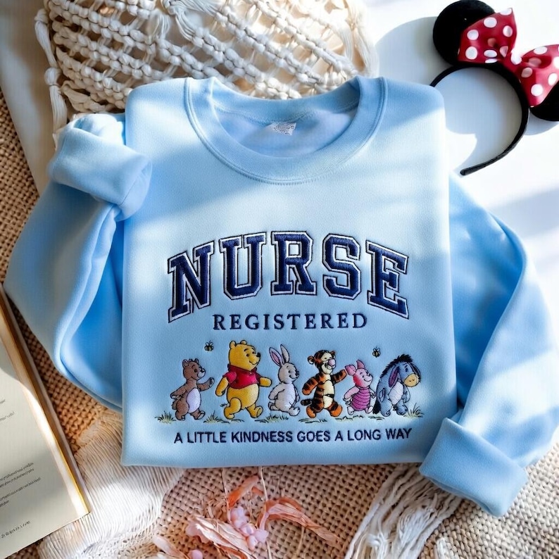 May include: Light blue sweatshirt with the word "NURSE" embroidered in navy blue, with "REGISTERED" below. Features cartoon characters and the phrase "A LITTLE KINDNESS GOES A LONG WAY". A Minnie Mouse ear headband is in the background.
