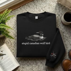 May include: Black sweatshirt featuring a loon illustration and the text "stupid canadian wolf bird." The sleeve has a hockey-themed design with the numbers 24 and 81. The sweatshirt is made of a soft material.