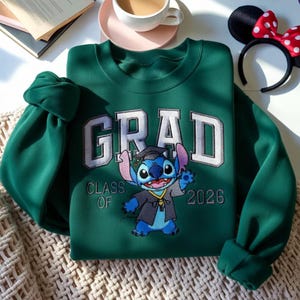Puede incluir: Sudadera verde oscuro con la palabra "GRAD" bordada en blanco y gris claro. Debajo, un personaje de dibujos animados con birrete y toga de graduaci&oacute;n. El texto "CLASS OF 2026" est&aacute; bordado debajo.