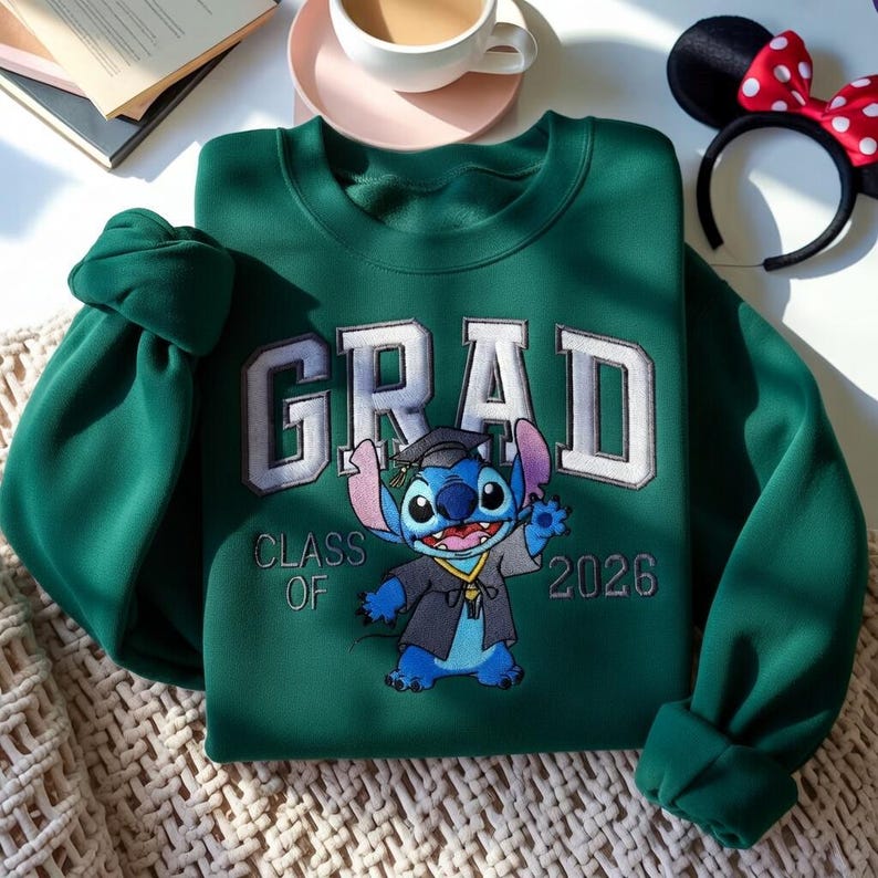 Puede incluir: Sudadera verde oscuro con la palabra "GRAD" bordada en blanco y gris claro. Debajo, un personaje de dibujos animados con birrete y toga de graduaci&oacute;n. El texto "CLASS OF 2026" est&aacute; bordado debajo.