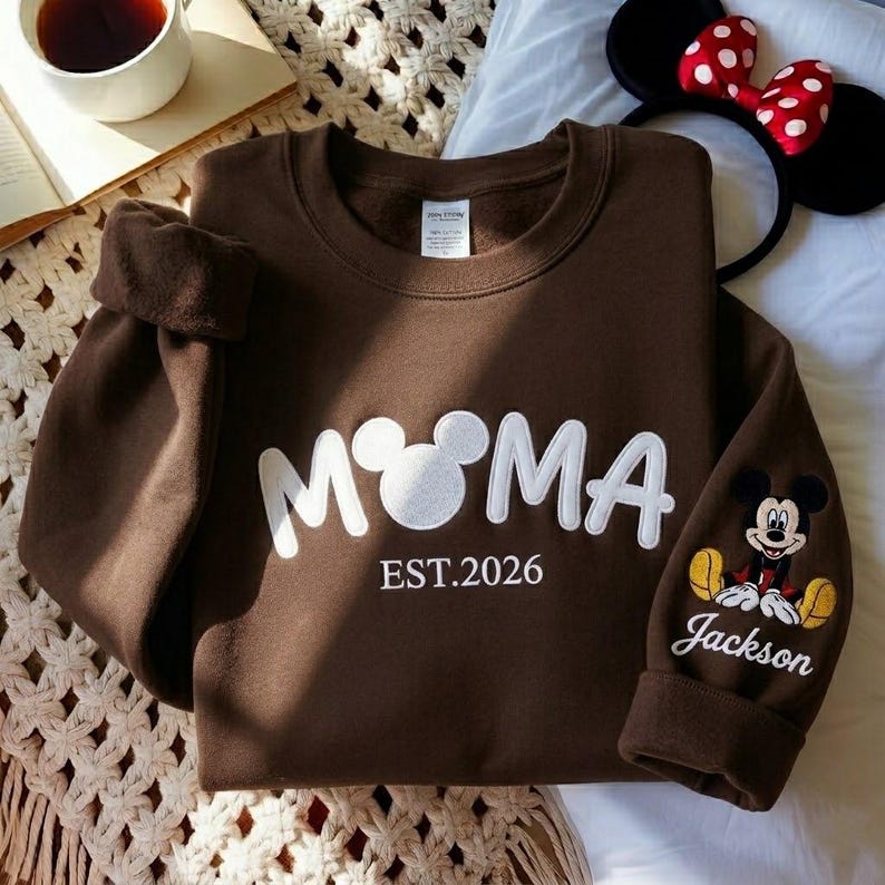 May include: A brown sweatshirt with the word "M♡MA" in white, featuring a Mickey Mouse head as the "O". The sleeve has a Mickey Mouse graphic and the name "Jackson". The text "EST.2026" is below the main text.