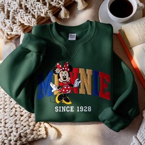 K&ouml;nnte beinhalten: Ein dunkelgr&uuml;nes Sweatshirt mit einem Minnie Mouse-Motiv. Das Wort "MINNIE" ist in bunten Blockbuchstaben dargestellt, darunter der Text "SINCE 1928". Die Cartoon-Figur Minnie Mouse tr&auml;gt ein rotes Polka-Dot-Kleid und eine Schleife.