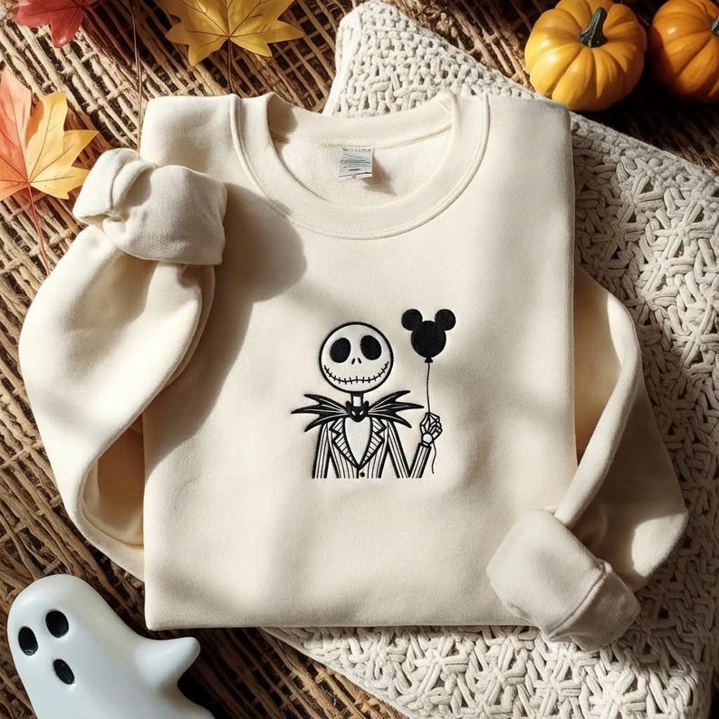 May include: A cream-colored crewneck sweatshirt featuring a black embroidered graphic of a skeletal character in a pinstriped suit holding a black Mickey Mouse-shaped balloon. The garment has long sleeves with ribbed cuffs and a ribbed neckline.