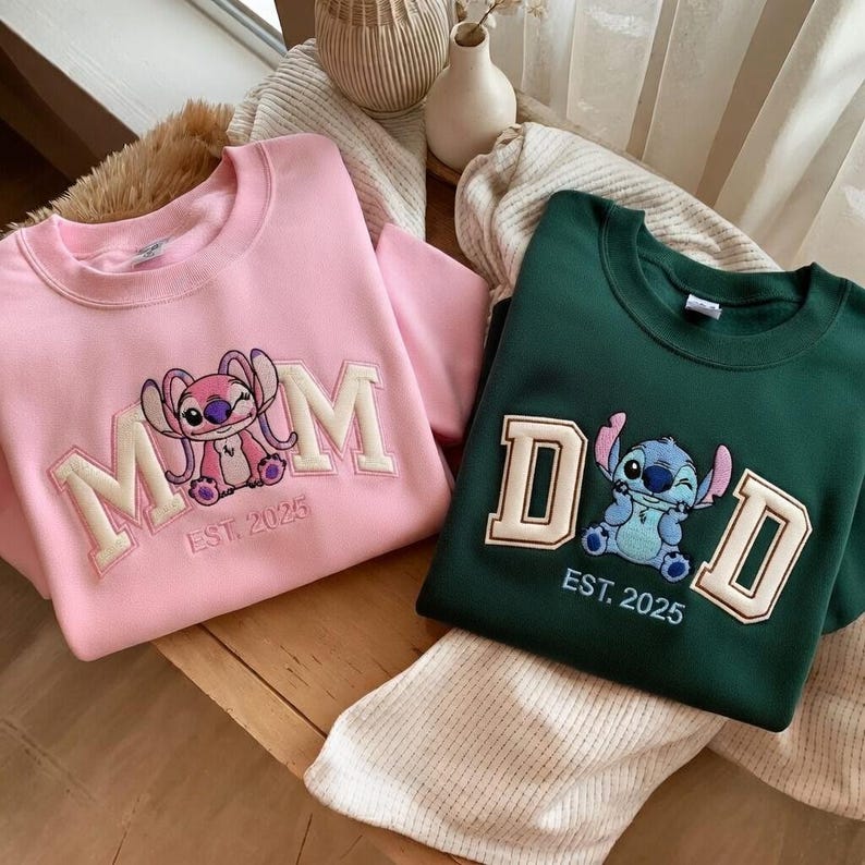 Puede incluir: Dos sudaderas, una rosa y otra verde oscuro, con dise&ntilde;os bordados. La sudadera rosa presenta la palabra "MOM" con un personaje de dibujos animados y "EST. 2025". La sudadera verde dice "DAD" con un personaje de dibujos animados y "EST. 2025".