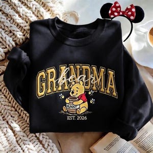 May include: Black sweatshirt with the word "GRANDMA" in gold block letters, with "bear" in cursive. A Winnie the Pooh graphic holding a honey pot is below the text, with "EST. 2026". A Minnie Mouse ear headband is in the background.