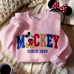 May include: A pale pink sweatshirt with the word "Mickey" embroidered in blue, red, and gold, and a cartoon Mickey Mouse. The text "SINCE 1928" is below. A headband with black ears and a red and white polka dot bow is also visible.