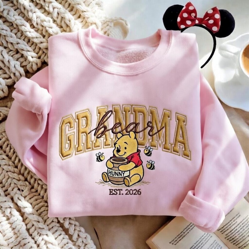May include: A light pink sweatshirt with the word "Grandma" in gold lettering and a Winnie the Pooh graphic. The graphic includes Winnie the Pooh holding a honey pot, bees, and the text "EST. 2026". A Minnie Mouse ear headband is visible.