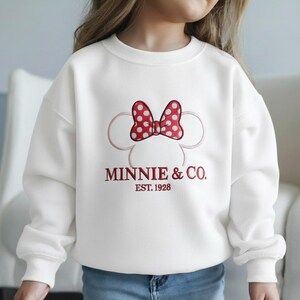 Op de afbeelding: Witte sweatshirt met een Minnie Mouse-ontwerp. Het ontwerp bevat een rode strik met witte stippen, Minnie Mouse-oren en de tekst "MINNIE & CO. EST. 1928" in het rood. Een comfortabele, casual top voor kinderen.