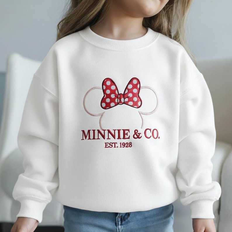 Op de afbeelding: Witte sweatshirt met een Minnie Mouse-ontwerp. Het ontwerp bevat een rode strik met witte stippen, Minnie Mouse-oren en de tekst "MINNIE & CO. EST. 1928" in het rood. Een comfortabele, casual top voor kinderen.