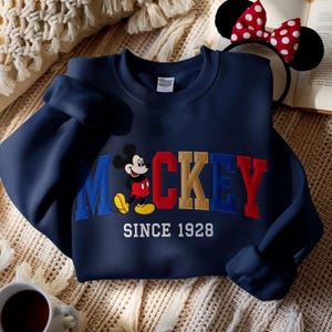 May include: Navy blue sweatshirt with the name "Mickey" embroidered in colourful letters, and a Mickey Mouse graphic. The text "SINCE 1928" is embroidered below. A pair of Mickey Mouse ears with a red and white polka dot bow is in the background.