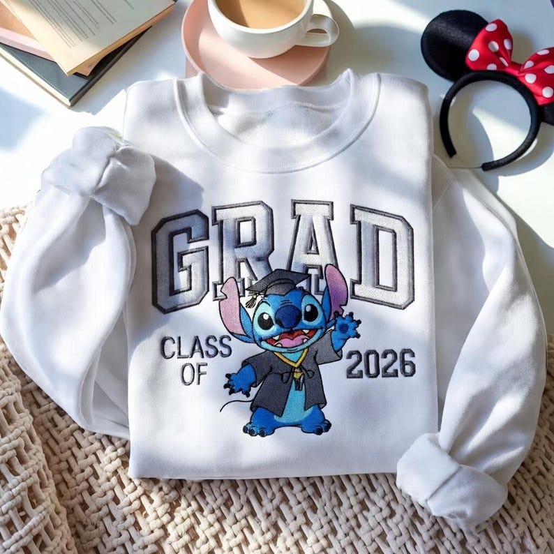 Puede incluir: Sudadera blanca con la palabra "GRAD" en letras grises y el texto "CLASS OF 2026". La sudadera presenta un personaje de dibujos animados con toga de graduaci&oacute;n. Se ve una diadema con orejas de Minnie Mouse.