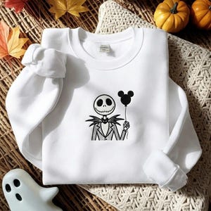 May include: A white long-sleeve crewneck sweatshirt featuring a black embroidered graphic of a skeleton character wearing a pinstriped suit and bowtie, holding a black balloon shaped like a mouse head in one hand, centered on the chest.