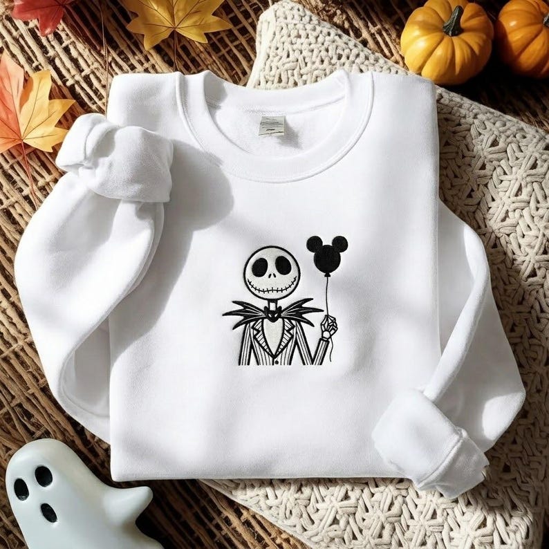 May include: A white long-sleeve crewneck sweatshirt featuring a black embroidered graphic of a skeleton character wearing a pinstriped suit and bowtie, holding a black balloon shaped like a mouse head in one hand, centered on the chest.