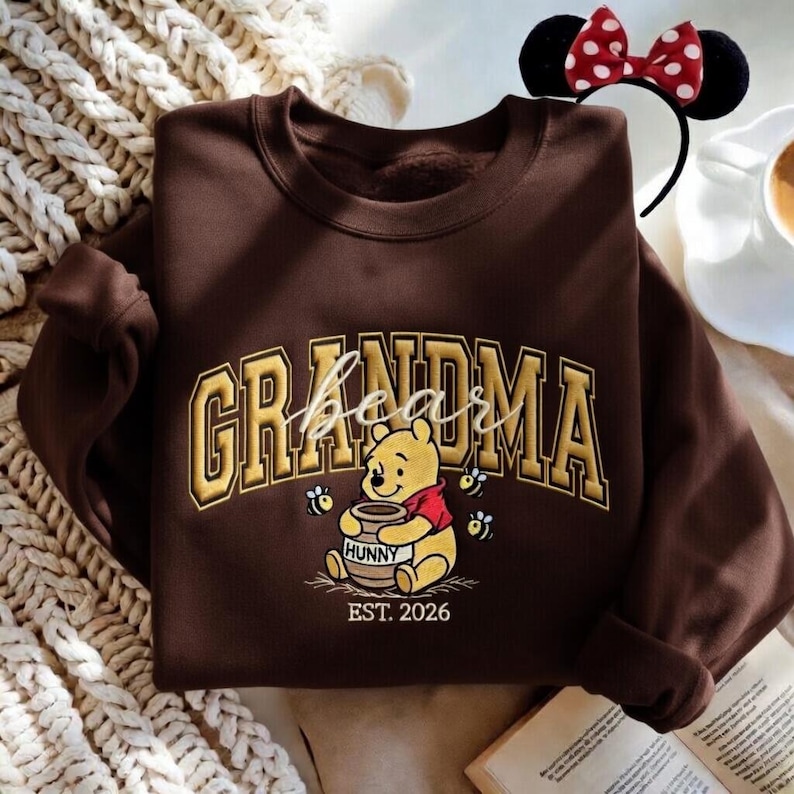 May include: A brown sweatshirt with the word "GRANDMA" in gold lettering and a Winnie the Pooh graphic. The graphic includes the word "bear" in script, a honey pot, bees, and the year "EST. 2026". A Minnie Mouse headband is in the background.