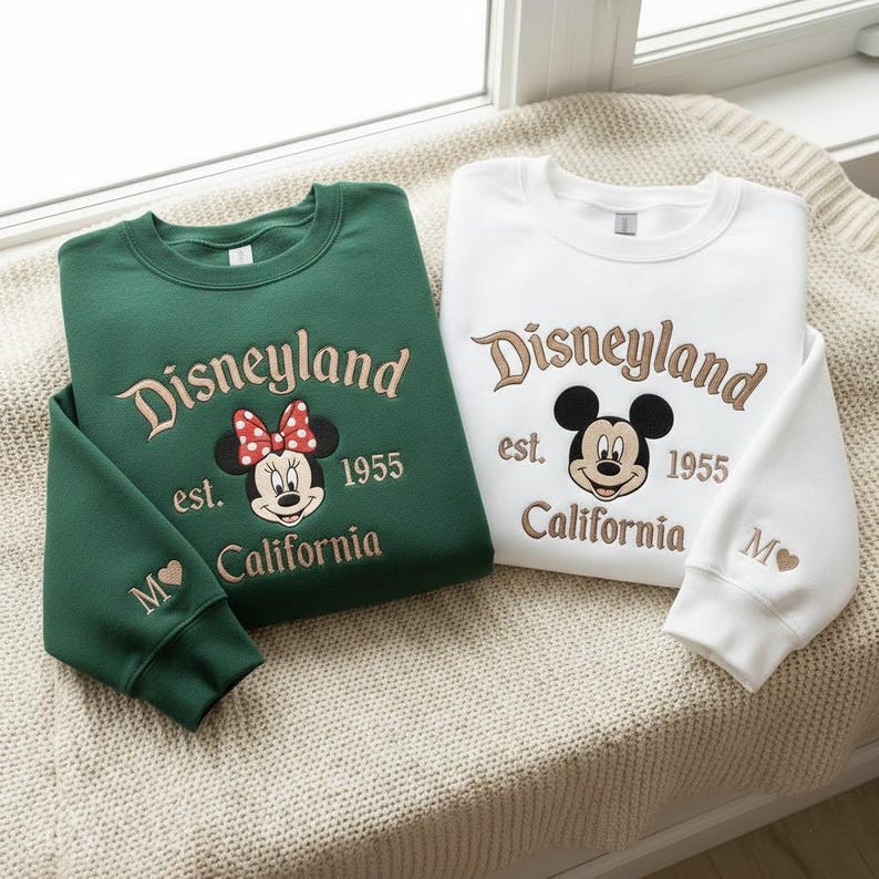 Puede incluir: Dos sudaderas, una verde y otra blanca, con bordado de Disneyland. La sudadera verde tiene un dise&ntilde;o de Minnie Mouse con un lazo rojo, la blanca Mickey Mouse. Ambas incluyen "est. 1955 California" y una letra "M" con un coraz&oacute;n.