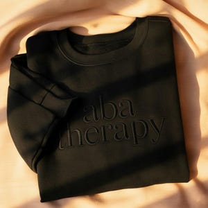 Può includere: Felpa nera girocollo con la scritta ricamata "aba therapy". La felpa è piegata e sembra essere realizzata in un materiale morbido. Lo sfondo è un tessuto color pesca.