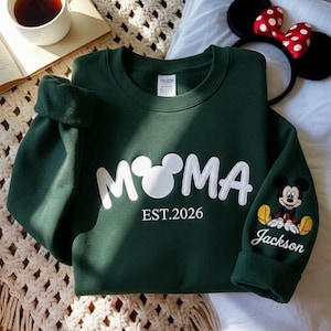 May include: A dark green sweatshirt with the word "M♡MA" in white, featuring a Mickey Mouse head as the "O". The sleeve has a Mickey Mouse graphic and the name "Jackson". The text "EST.2026" is below the main text.