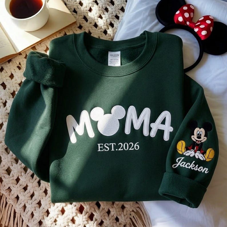 May include: A dark green sweatshirt with the word "M♡MA" in white, featuring a Mickey Mouse head as the "O". The sleeve has a Mickey Mouse graphic and the name "Jackson". The text "EST.2026" is below the main text.