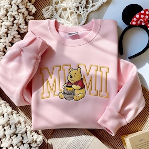以下が含まれることがあります： 金色の文字で「MIMI」と書かれた、くまのプーさんのイラストが描かれた薄いピンク色のスウェットシャツ。 漫画のキャラクターは蜂蜜の壺を持っています。 ミニーマウスの耳と赤い水玉模様のリボンが付いた黒いヘアバンドが見えます。