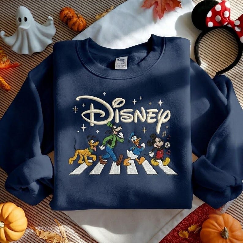 Peut inclure: Un sweat-shirt bleu marine &agrave; col rond avec une broderie repr&eacute;sentant Pluto, Dingo, Donald et Mickey marchant sur un passage pi&eacute;ton blanc. Au-dessus des personnages, le mot Disney est brod&eacute; dans une police cursive de couleur cr&egrave;me.